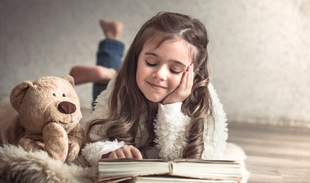 little girl reading a book with a teddy bear on the floor, concept of relaxation and friendship